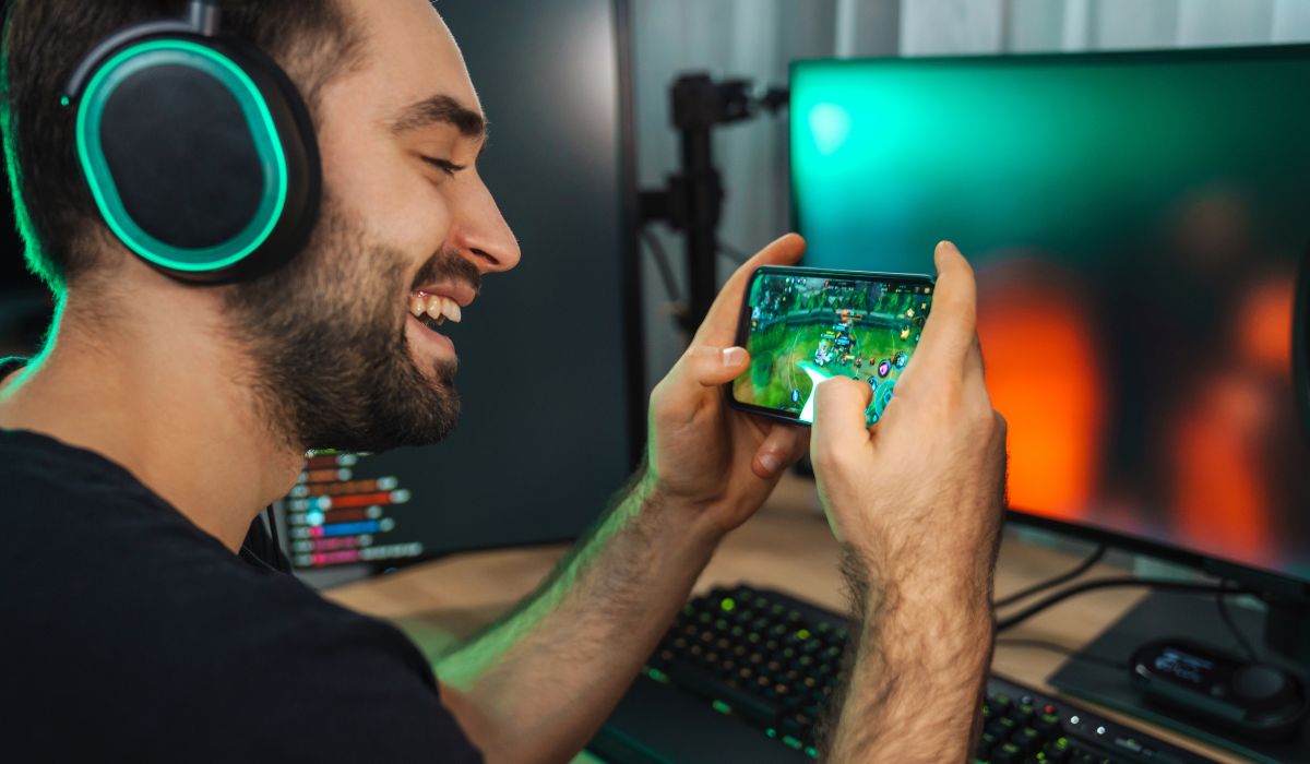 Un hombre está jugando un juego móvil en su teléfono junto a una computadora.