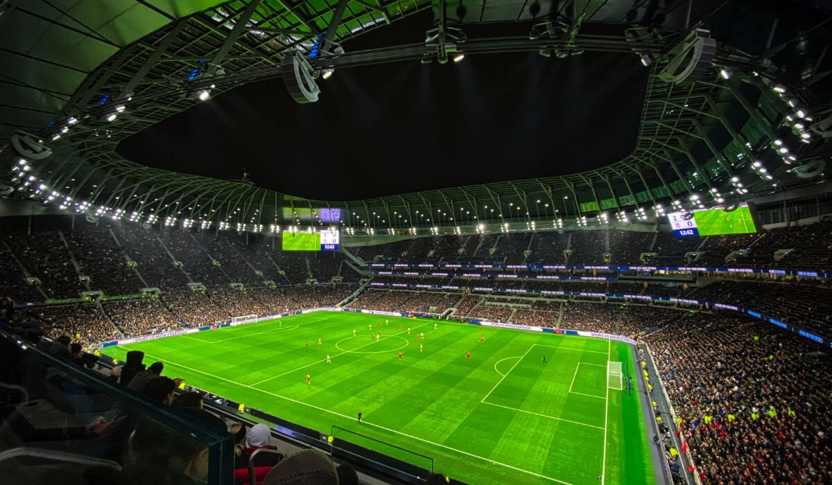 Una vista nocturna de un estadio de fútbol lleno de espectadores, mostrando un partido en desarrollo en el campo bien iluminado. El marcador y las pantallas grandes son visibles y muestran los detalles del juego junto con publicidad dentro del fútbol.