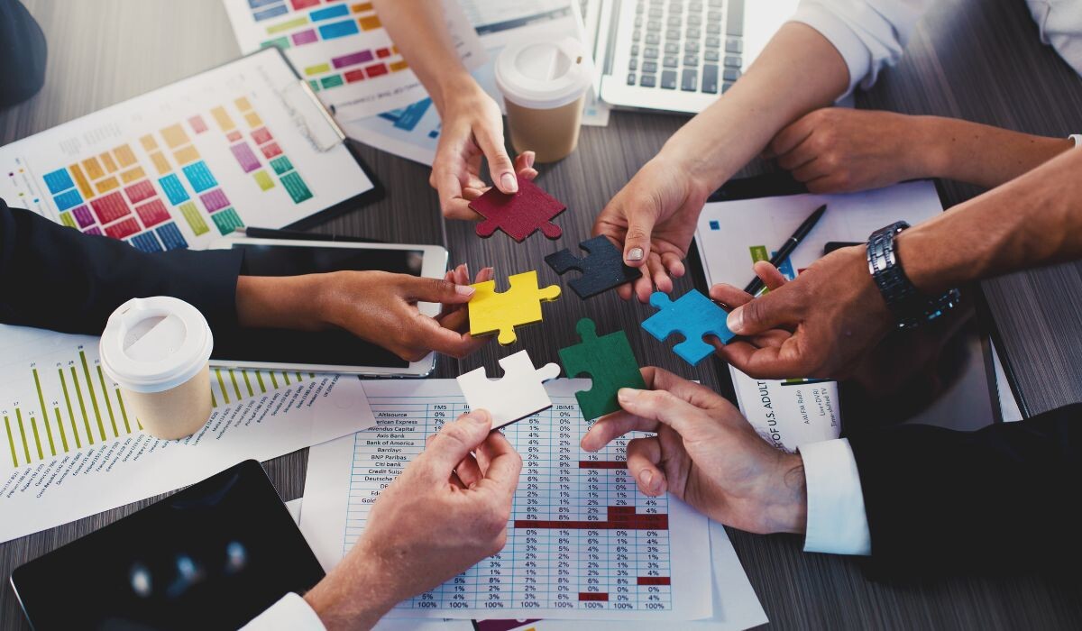 Un grupo de personas realiza trabajo colaborativo alrededor de una mesa, ensamblando coloridas piezas de un rompecabezas. Sobre la mesa hay gráficos, un ordenador portátil, tazas de café y papeles.