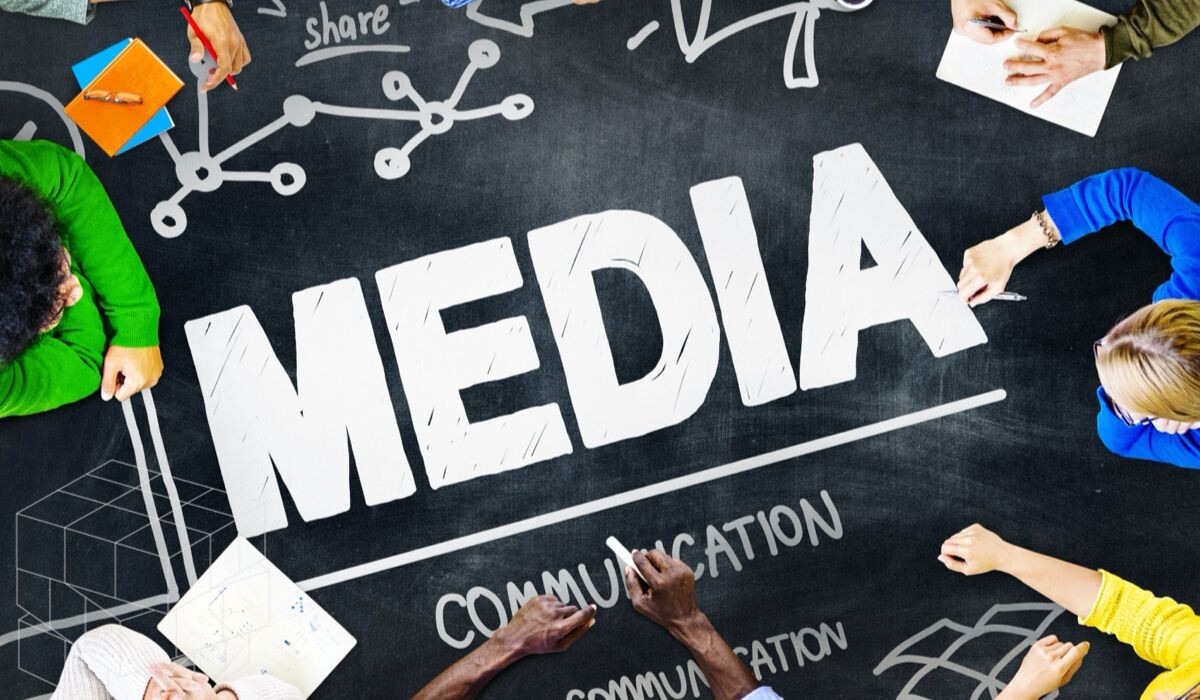 Un grupo de personas se sientan alrededor de una pizarra con la palabra "MEDIA" escrita en letras grandes; la pizarra incluye varios diagramas y dibujos relacionados con la comunicación, destacando su estrategia de medios.