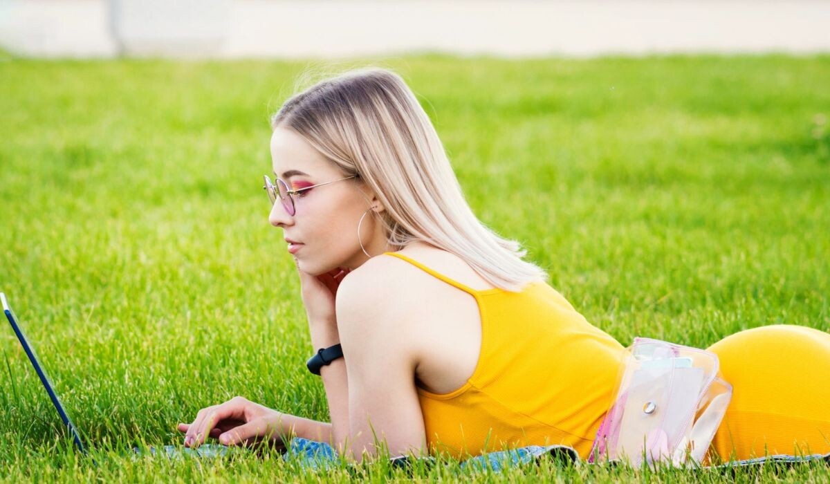 Una mujer vestida de amarillo yace boca abajo sobre el césped, trabajando como escritora independiente en su computadora portátil. Lleva gafas y tiene una bolsa transparente a su lado.