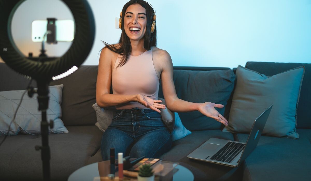 Una persona con cabello largo y auriculares sonríe mientras está sentada en un sofá. Hacen un gesto hacia una computadora portátil, con un anillo de luz y varios elementos sobre una mesa frente a ellos, encarnando la esencia de los influencers y la publicidad.