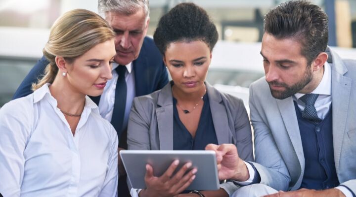 Cuatro profesionales de negocios están reunidos alrededor de una tableta en una oficina, discutiendo atentamente su contenido mientras revisan su página de empresa de LinkedIn.