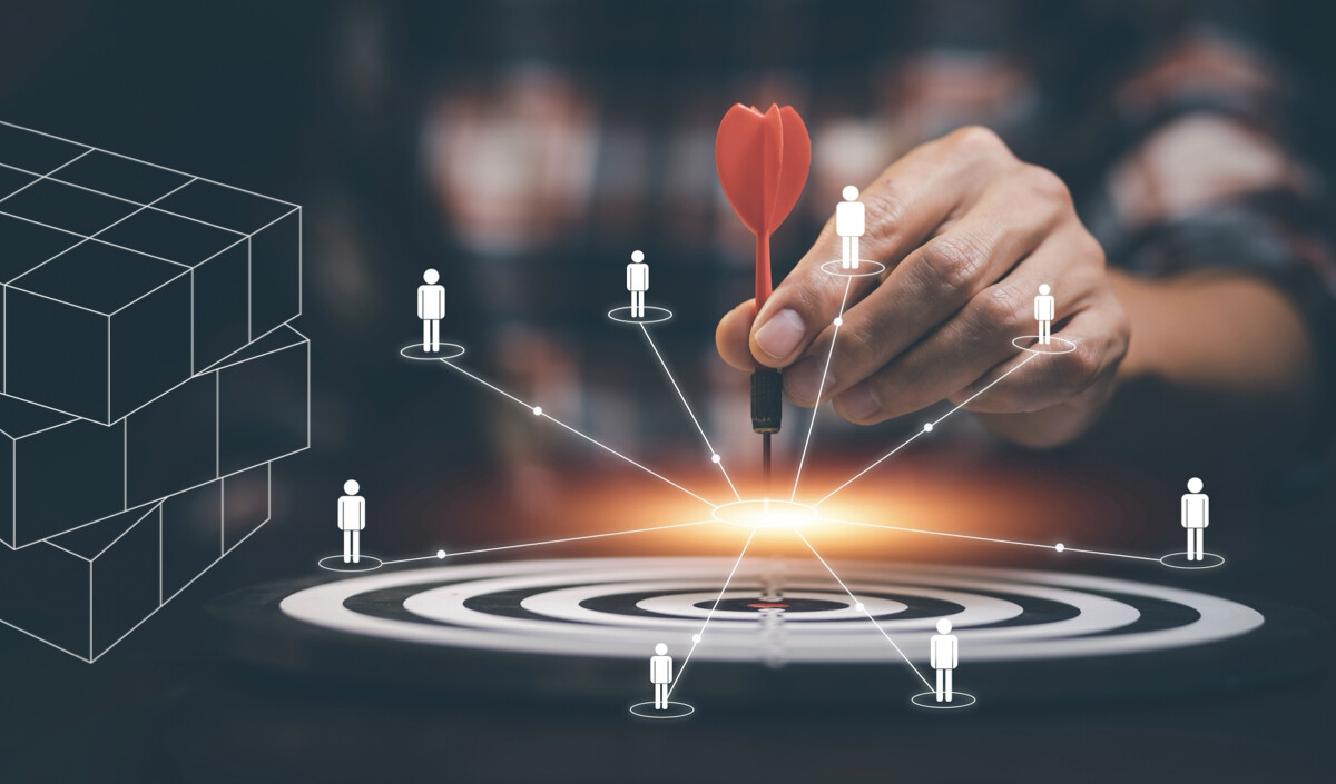 Una mano coloca un dardo rojo en el centro de una diana. Alrededor del tablero hay figuras humanas ilustradas conectadas por líneas, que representan grupos objetivos. Un gráfico de cubo está en el lado izquierdo.