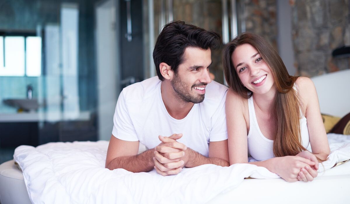 Un hombre y una mujer, ambos vestidos con camisas blancas, yacen en una cama, sonriendo y mirándose con sexualidad positiva en un dormitorio bien iluminado.