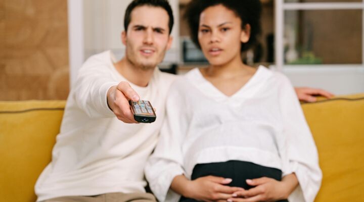Una pareja está sentada en un sofá amarillo, el hombre sostiene un control remoto y la mujer apoya las manos sobre su vientre de embarazada. Mientras miran la televisión, no pueden evitar reírse de la publicidad engañosa de los llamativos anuncios.