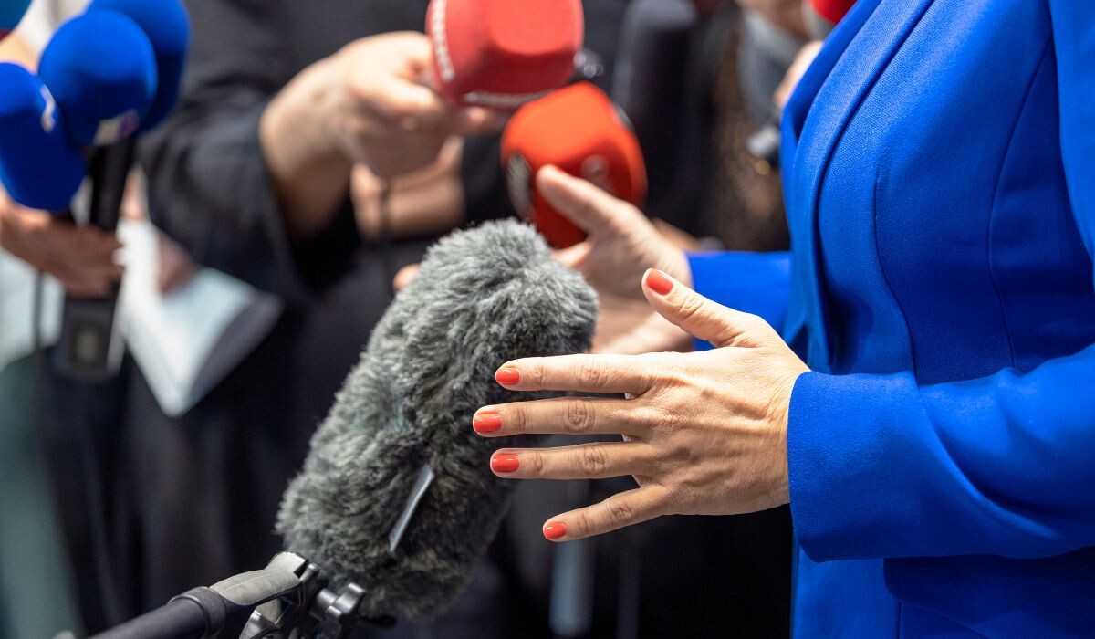 Una persona con traje azul, experta en relaciones públicas, habla con seguridad ante un grupo de periodistas que sostienen micrófonos.