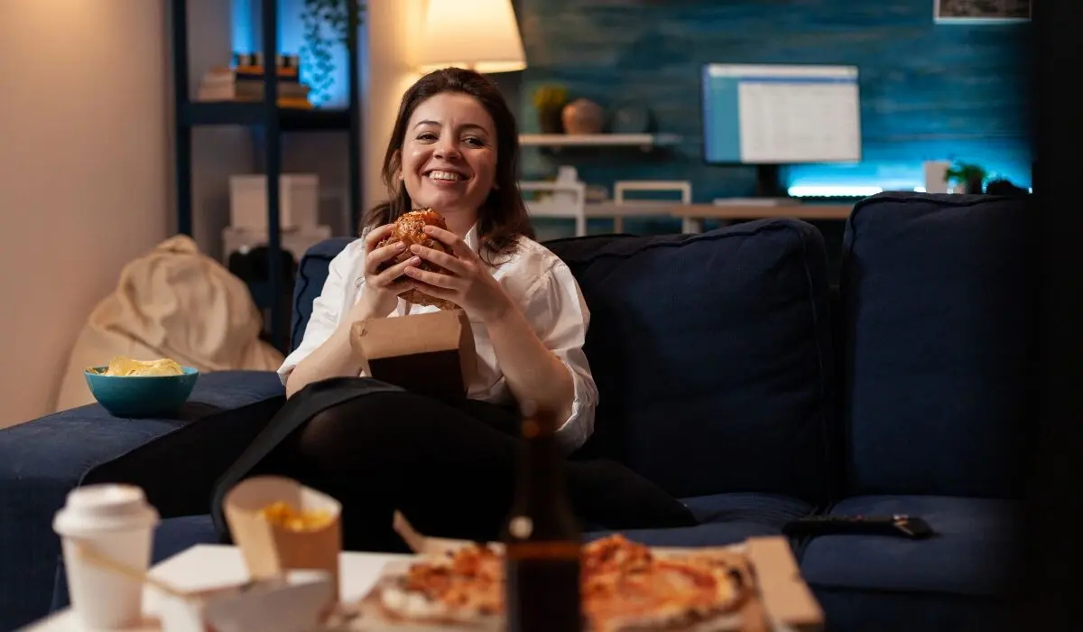 Una mujer está sentada en un sofá, sonriendo y comiendo una hamburguesa mientras mira compulsivamente su serie favorita. En una mesa frente a ella hay una pizza, una bebida y un plato de papas fritas. La cálida iluminación de la habitación y la pared con acentos azules crean el telón de fondo perfecto para esta relajada escena de marketing.