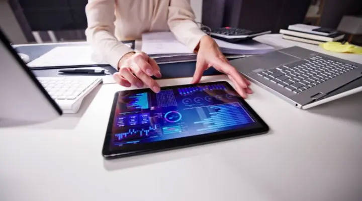 En medio de un mar de documentos de oficina y una computadora portátil, una persona interactúa con una tableta que muestra gráficos y cuadros financieros, adentrándose en predicciones tecnológicas para navegar por el panorama digital.
