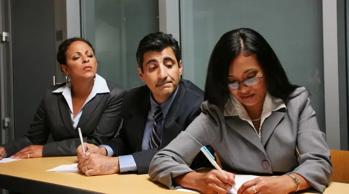 Tres personas vestidas de negocios están sentadas a una mesa. La mujer de la izquierda y el hombre del medio miran a la mujer de la derecha, que está escribiendo pensativamente en un papel; su concentración ejemplifica una fuerte marca personal.