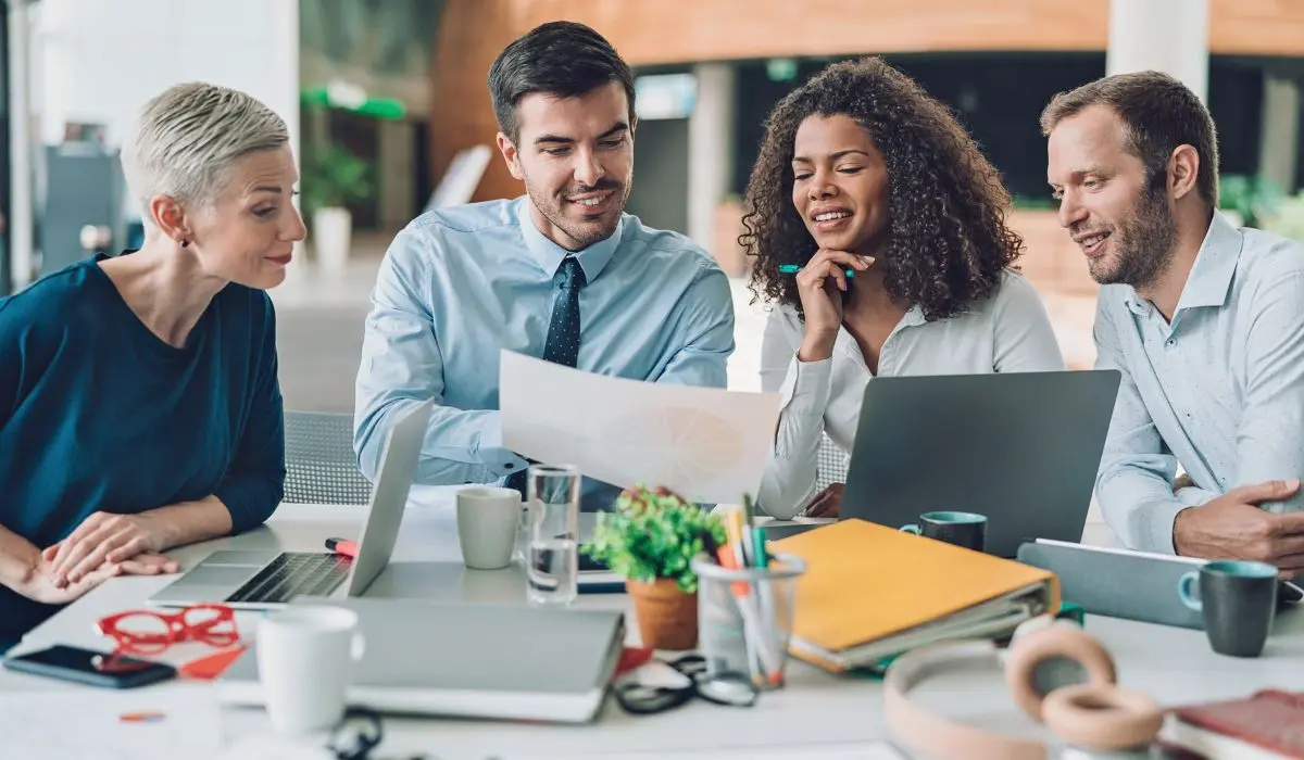 Cuatro colegas están sentados en una mesa con ordenadores portátiles y documentos, participando en una discusión.