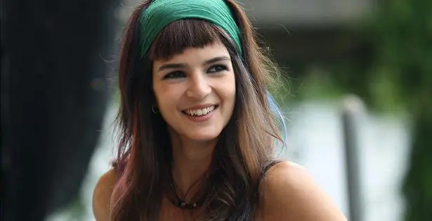Una persona con cabello largo, que lleva una diadema verde y sonríe, mira hacia un lado.