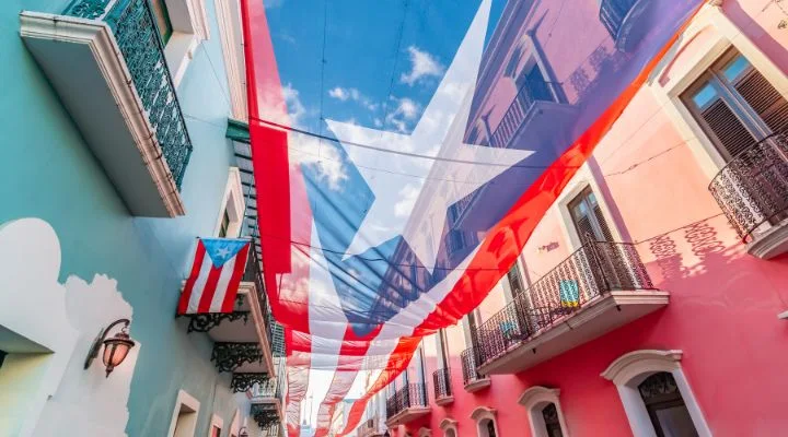 La imagen muestra una gran bandera puertorriqueña que cuelga entre las Residencias, ubicadas a lo largo de una calle angosta. Desde los balcones se puede ver la escena, donde también se ve una bandera más pequeña.