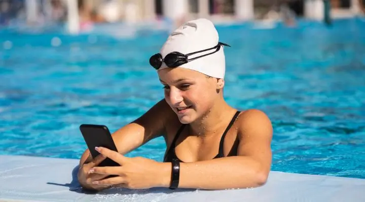 Una persona con gorro de baño y gafas se apoya casualmente en el borde de la piscina, usando un teléfono inteligente para interactuar con chatbots conversacionales.