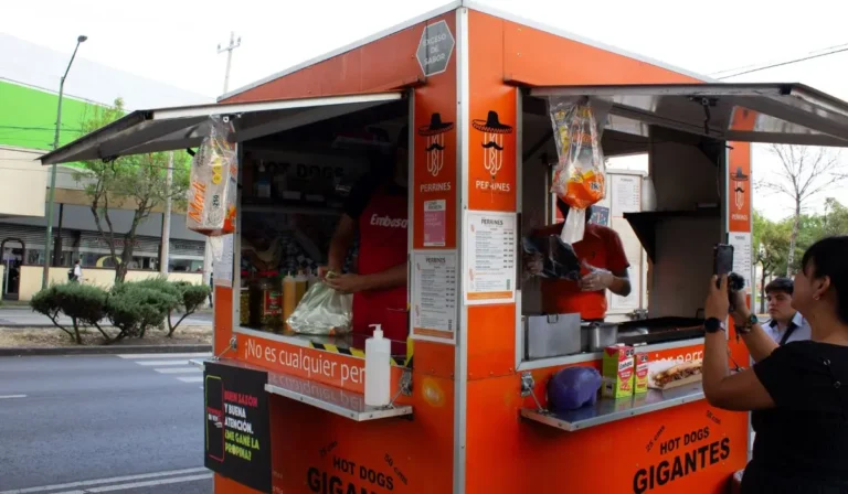 Una persona hace un pedido de puestos de comida naranja de perritos calientes en una calle concurrida. El puesto, un ejemplo vibrante de "Puestos de comida", muestra un menú y un cartel que proclama "Hot Dogs Gigantes". Cerca, otra persona parece estar capturando el momento con una foto.