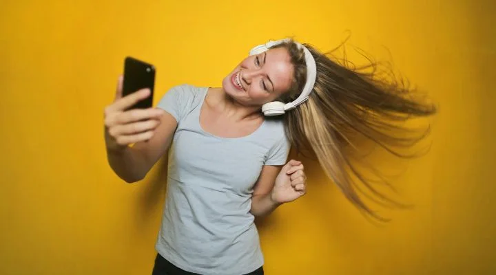 Una persona con auriculares, inmersa en su proceso creativo, se toma una selfie mientras su cabello se mueve suavemente. Está de pie frente a un fondo amarillo vibrante y viste una camisa gris claro.