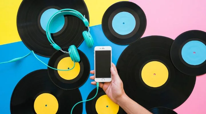 Discos de vinilo y auriculares turquesa se encuentran dispersos sobre un fondo colorido, creando un vibrante santuario para los fans de Taylor Swift. Una mano sostiene un teléfono inteligente, conectado expertamente a los auriculares, listo para reproducir los últimos éxitos de la icónica cantante.