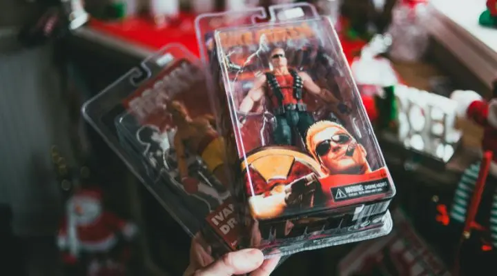 En una mano, dos figuras de acción en envases de plástico capturan la esencia de la alegría navideña. Una de ellas presenta a un personaje musculoso con gafas de sol y una camiseta roja sin mangas, deleitando a los kidults entre las decoraciones festivas de fondo.