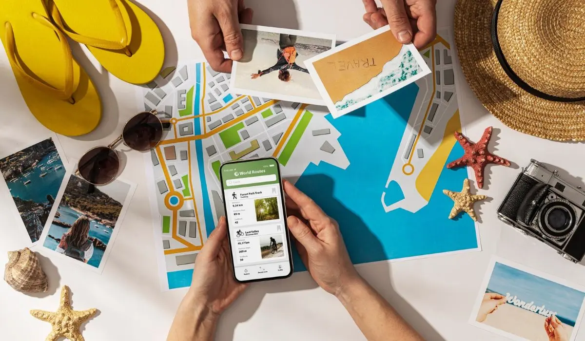 Manos sosteniendo un smartphone mostrando planes de vacaciones sobre un mapa. Alrededor hay fotos, un sombrero, sandalias, conchas, estrellas de mar y una cámara.
