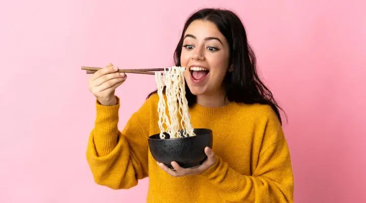 Una mujer con un suéter amarillo sostiene palillos y un tazón, comiendo fideos sobre un fondo rosa comiendo como símbolo del marketing emocional 