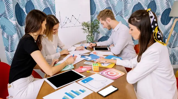 Cuatro personas del departamento de marketing se sientan en una mesa con tablets e iconos de redes sociales, discutiendo estrategias y anotando ideas. Un gráfico en la pared, detrás de ellas, complementa su análisis.