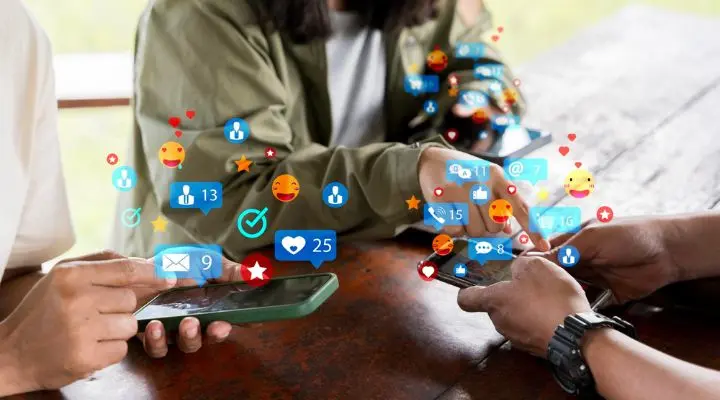 Tres personas están sentadas en una mesa usando teléfonos inteligentes, generando sin problemas diversas notificaciones digitales como me gusta, mensajes y emojis, simbolizando la conectividad y mostrando las tendencias en social media