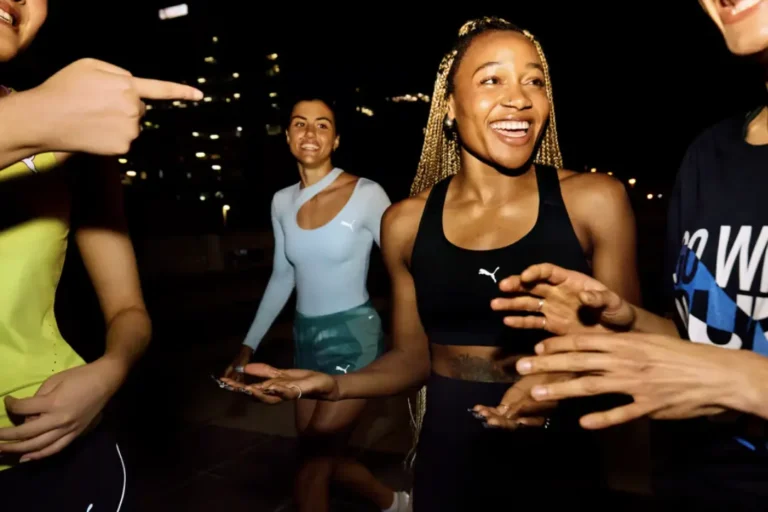 Cuatro mujeres vestidas con ropa deportiva de PUMA, sonriendo e interactuando al aire libre por la noche con las luces de la ciudad de fondo: una escena vibrante que captura la esencia del marketing deportivo.