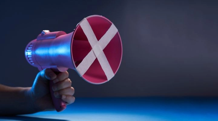 Una mano sostiene un megáfono rosa con una X blanca hecha de cinta que cubre el altavoz, simbolizando la censura, sobre un fondo oscuro.