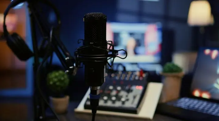 Un primer plano de un micrófono con un filtro antipop en un estudio de grabación, perfecto para podcasting, con auriculares, una consola de mezclas y pantallas de computadora borrosas en el fondo.