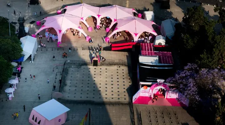 Desde una perspectiva aérea, la vibrante plaza exhibe tiendas y puestos Sedal con techos rosados, enclavados entre frondosos árboles y senderos sinuosos. La gente se reúne bajo y alrededor de estos acogedores refugios.