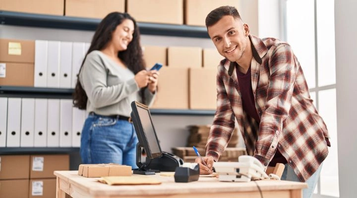 Hombre con camisa a cuadros escribiendo en un escritorio con paquetes y una computadora; mujer en el fondo usando un teléfono cerca de estantes con cajas y carpetas mostrando el emprendimiento en México