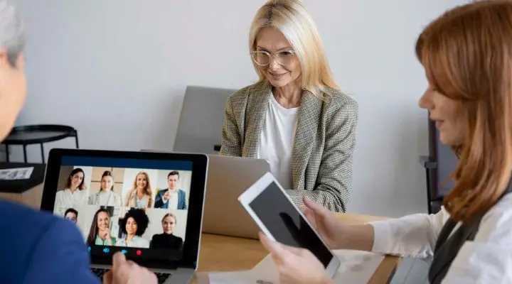Tres mujeres trabajan en Relaciones públicas: una usa una computadora portátil, otra sostiene una tableta y una tercera participa en una videollamada centrada en la ética con varias personas en la pantalla de su computadora portátil.