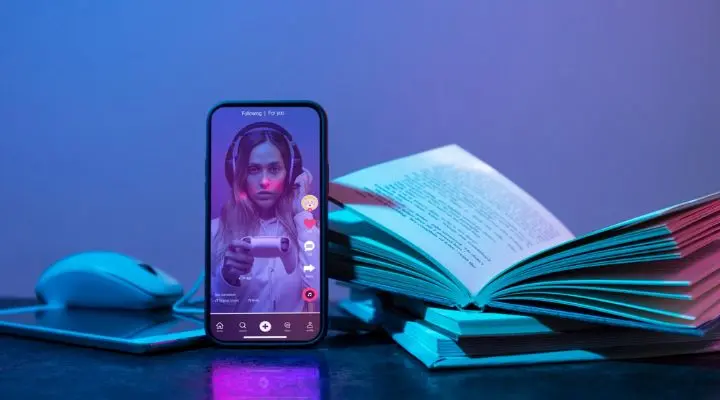 Un teléfono inteligente que muestra un video de TikTok se encuentra en posición vertical sobre una mesa junto a un libro abierto y un mouse inalámbrico, todo bañado por una vibrante iluminación rosa y azul.