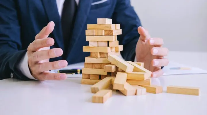 Una persona con traje hace un gesto hacia una torre de bloques de madera derribada sobre una mesa blanca; los bloques dispersos simbolizan los riesgos y desafíos de la compra de medios.