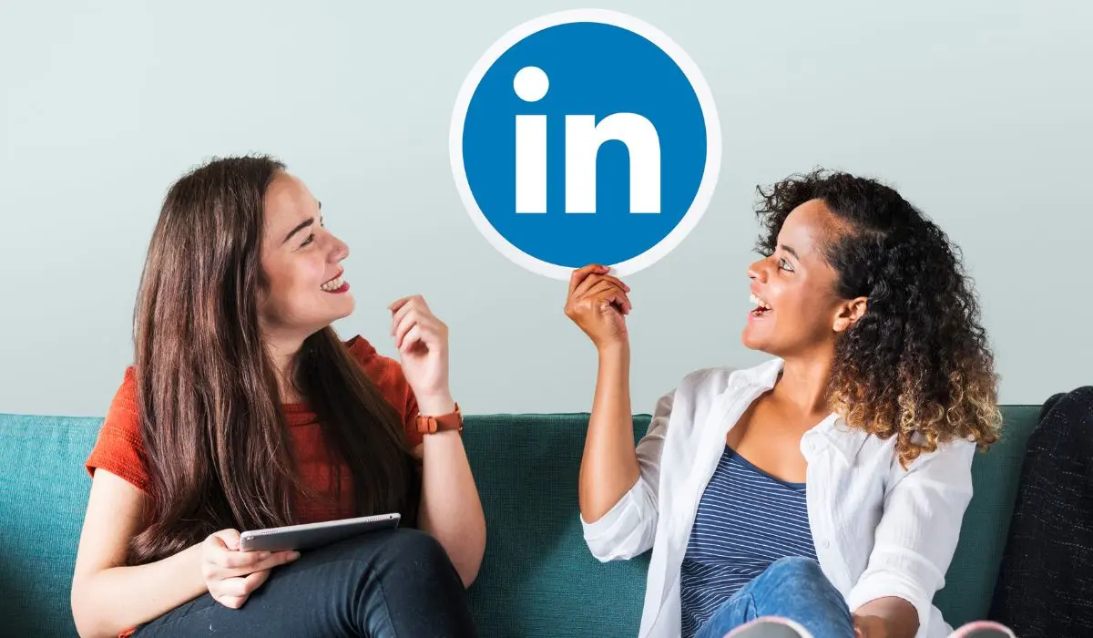 Dos mujeres sentadas en un sofá sonriendo; una sostiene una tableta mientras la otra muestra con orgullo un cartel con el logotipo de LinkedIn, celebrando su comunidad profesional.