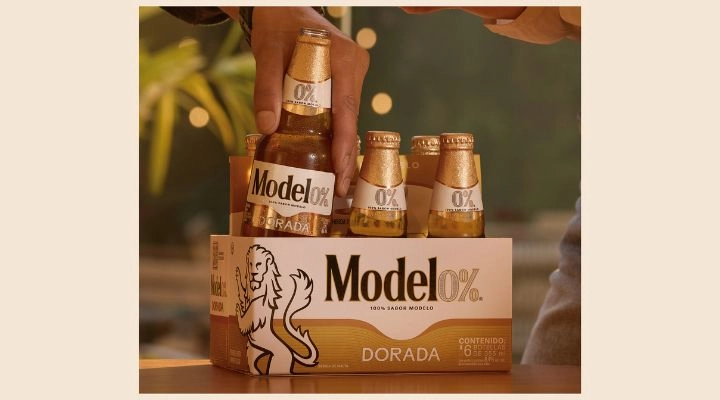 Una mano levanta una botella de un pack de seis de cerveza sin alcohol model0% Dorada sobre una mesa.
