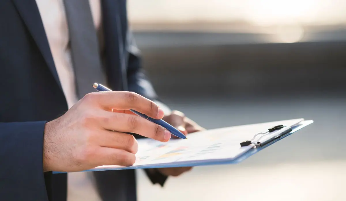 Una persona con traje sostiene un bolígrafo y señala un papel con gráficos en un portapapeles.