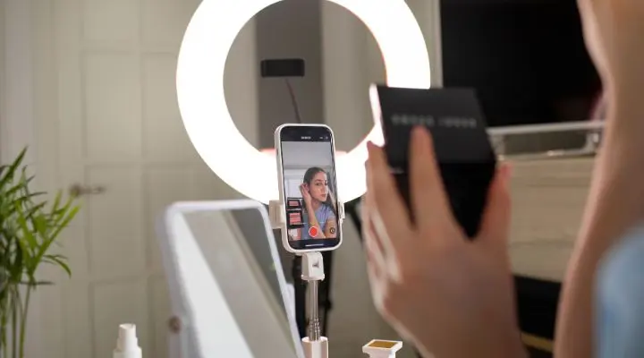 Persona grabando un tutorial de maquillaje en un teléfono inteligente, colocada frente a un anillo de luz, mostrando estrategias de video mientras sostiene un producto de maquillaje con un espejo visible en el fondo.