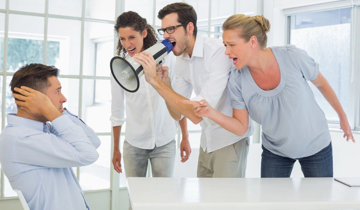 Tres personas se enfrentan en voz alta a un hombre sentado en un escritorio: uno grita a través de un megáfono sobre RRPP, mientras se tapa los oídos en señal de frustración.