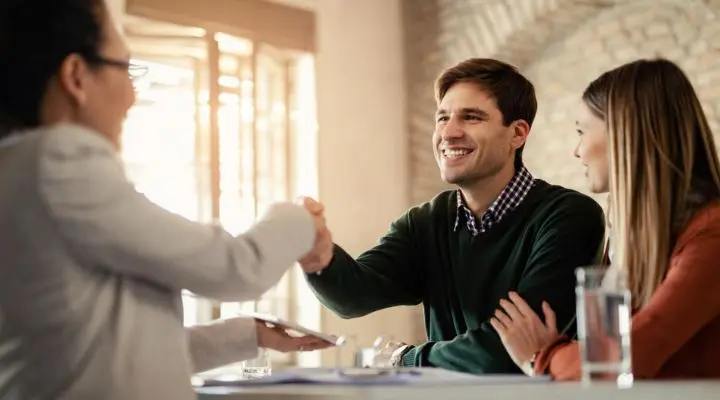 Un hombre y una mujer se sientan en una mesa, sonriendo, mientras el hombre le estrecha la mano a otra persona frente a ellos en un ambiente interior luminoso, sellando un trato de ventas exitoso.