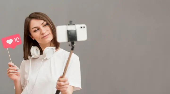 Una mujer con auriculares alrededor del cuello se toma una selfie con un teléfono inteligente en un palo para selfies, sosteniendo un cartel que muestra un corazón y el número 10, capturando la esencia de los influenciadores en acción.