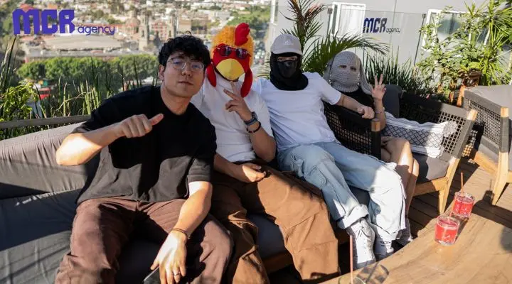 Cuatro personas están sentadas en un sofá del patio al aire libre; tres creadores de contenido usan máscaras, incluida una máscara de pollo y una máscara de malla, mientras que la persona de la izquierda no tiene máscara y levanta un dedo.