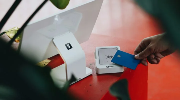 Una persona sostiene una tarjeta azul sin contacto cerca de una terminal de pago en un mostrador rojo, capturando el auge de los neobancos en México, con un monitor blanco en el fondo y algunas hojas verdes parcialmente visibles.